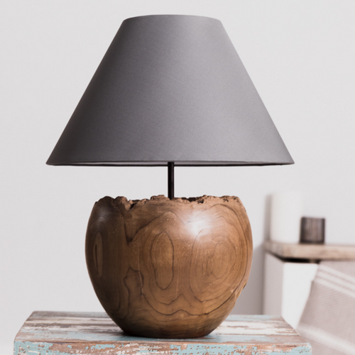 A rustic wood table lamp with a solid teak wood base and a gray conical lampshade, displayed on a wooden table.