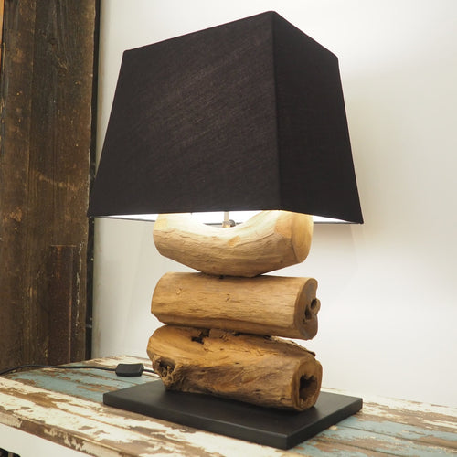 A wooden desk lamp with a rustic teak root base, featuring three chunky pieces stacked together. The lamp is topped with a black square lampshade, and it's placed on a wooden surface with a dark background.