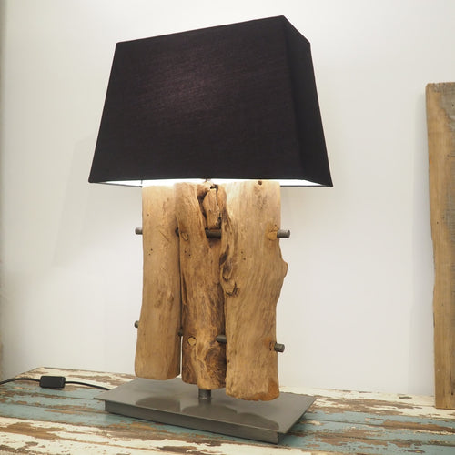 A wooden table lamp base featuring 3 pieces of natural wood connected on a metal frame with a metal base, positioned on a wooden surface with a black shade on top.