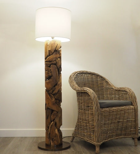 A floor lamp with a teak wood base and shaft, with a natural cotton colour  lamp shade on top, standing next to a wicker chair.